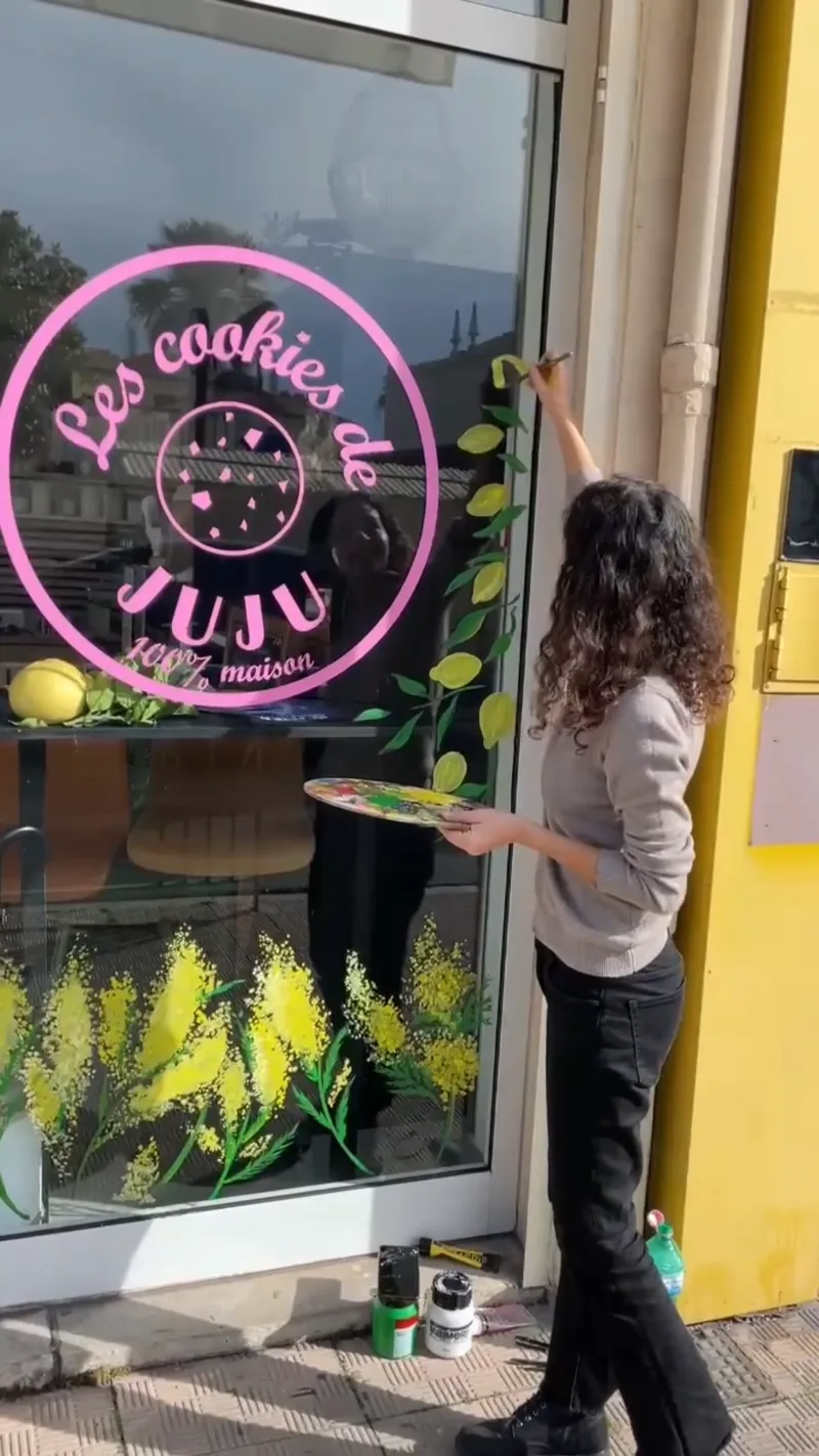 Example of a hand-painted logo by Giulia Dal Bon: she paints a shop’s pink logo on its window, framed by mimosa.