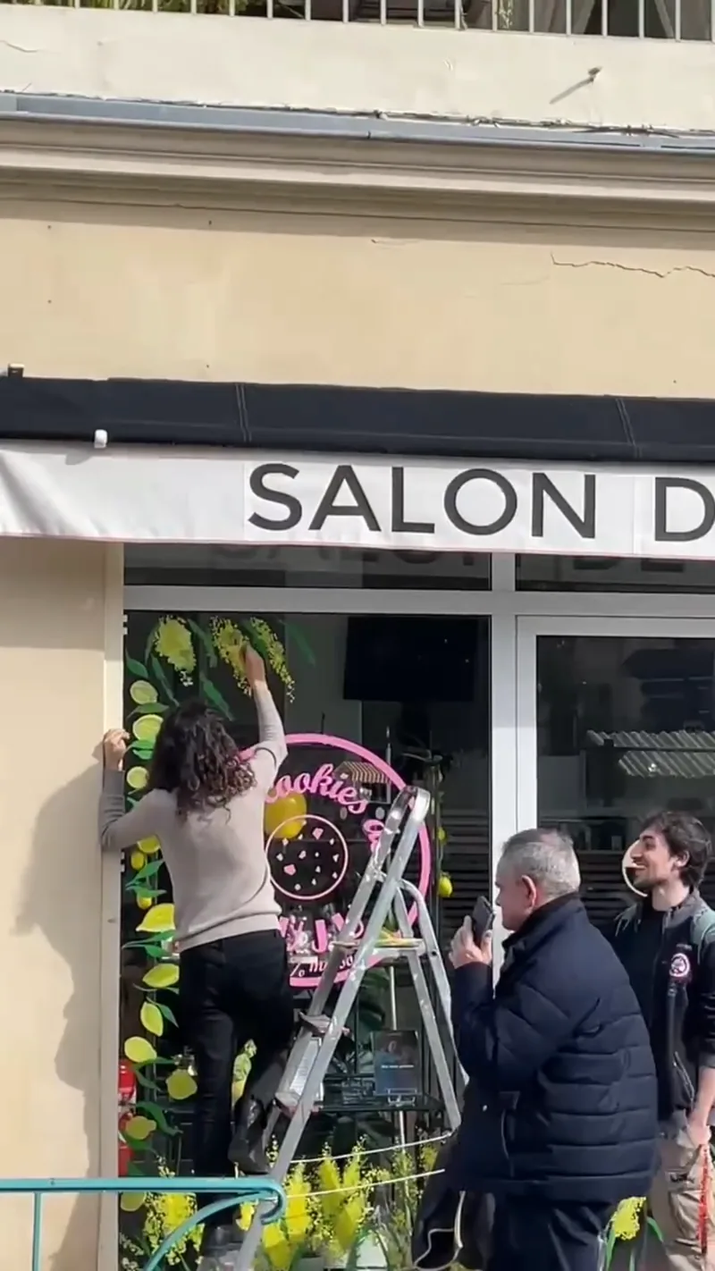 Giulia Dal Bon peint la vitrine d’une boutique pendant que des passants la regardent et la photographient.