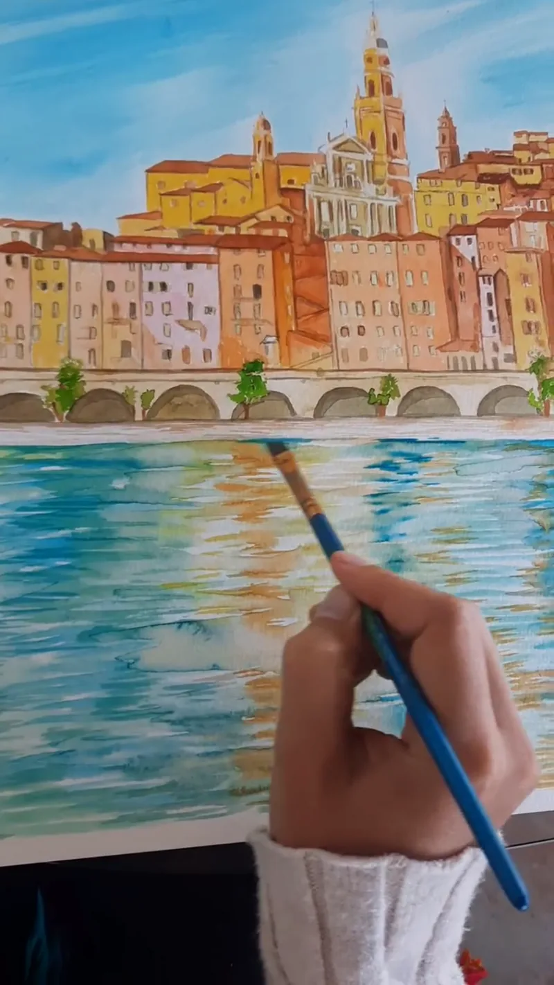 Giulia Dal Bon peint au pinceau les reflets d’une ville ocre et dorée dans l’eau qui la borde.