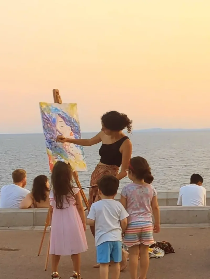 In riva al mare, tre bambini guardano Giulia Dal Bon dipingere dal vivo un volto di donna dai capelli blu.