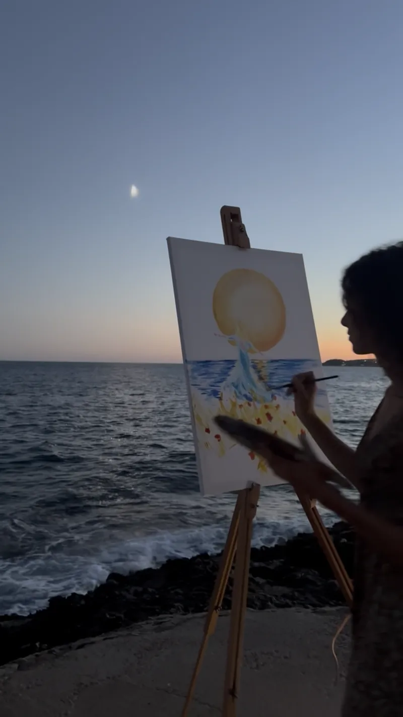 Au crépuscule, sous un croissant de lune, Giulia Dal Bon peint au chevalet face à la mer.