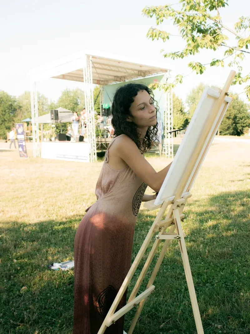 Dans un parc, Giulia Dal Bon peint au chevalet devant une scène dressée en plein air.