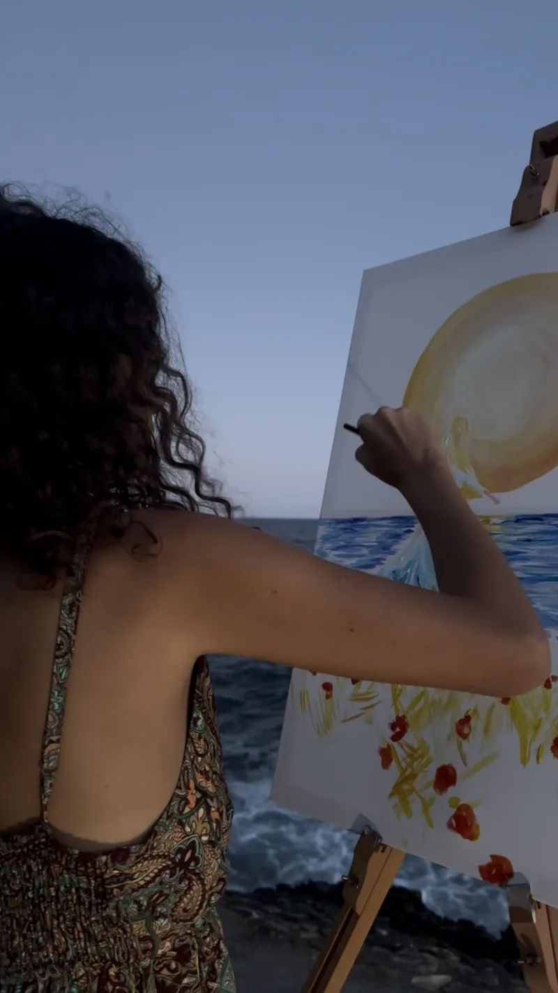 Tout près de la toile, Giulia Dal Bon peint le reflet du soleil et les fleurs rouges, face à la mer au crépuscule.