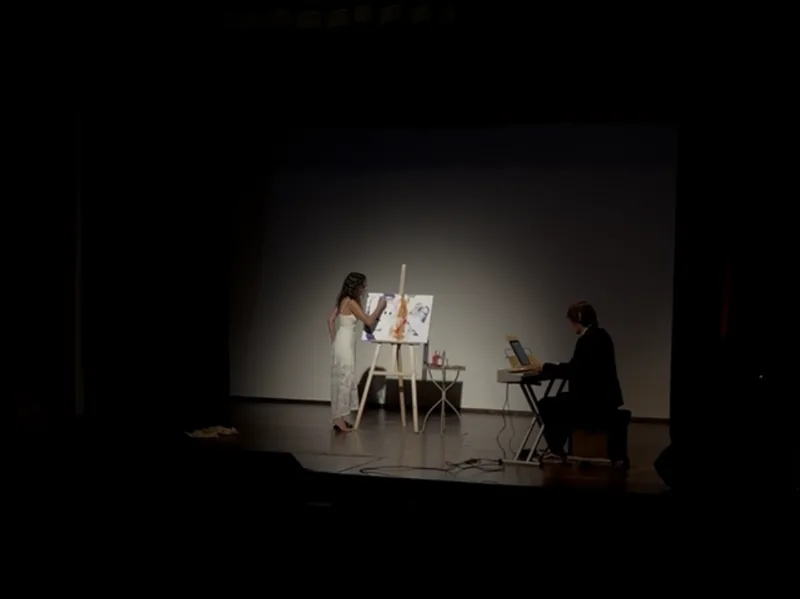 Spettacolo piano e pittura: su un palco di teatro immerso nel buio, Giulia Dal Bon dipinge nella luce, Orlando alla tastiera.