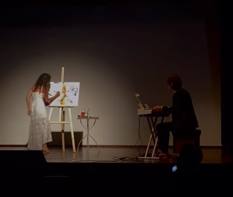 Sur une scène de théâtre, Giulia Dal Bon peint au chevalet dans la lumière des projecteurs pendant qu’Orlando joue au clavier.