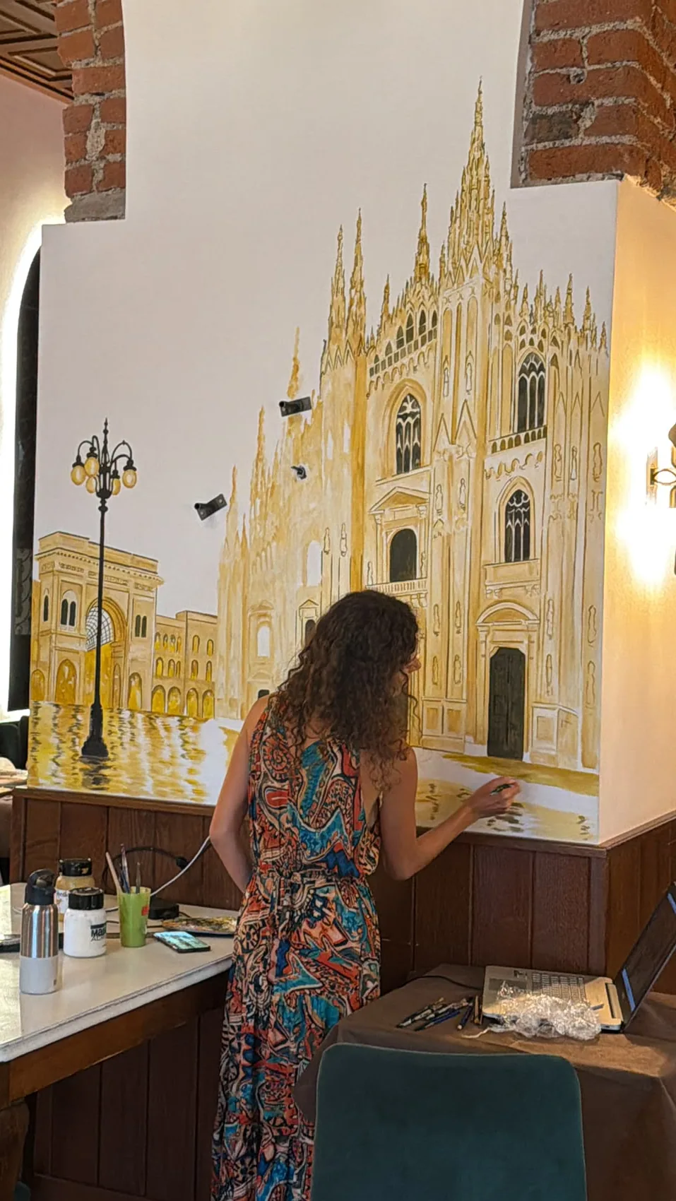 Il disegno in ocra del murale copre tutta la parete, e Giulia Dal Bon comincia a dipingere la piazza ai piedi del Duomo.