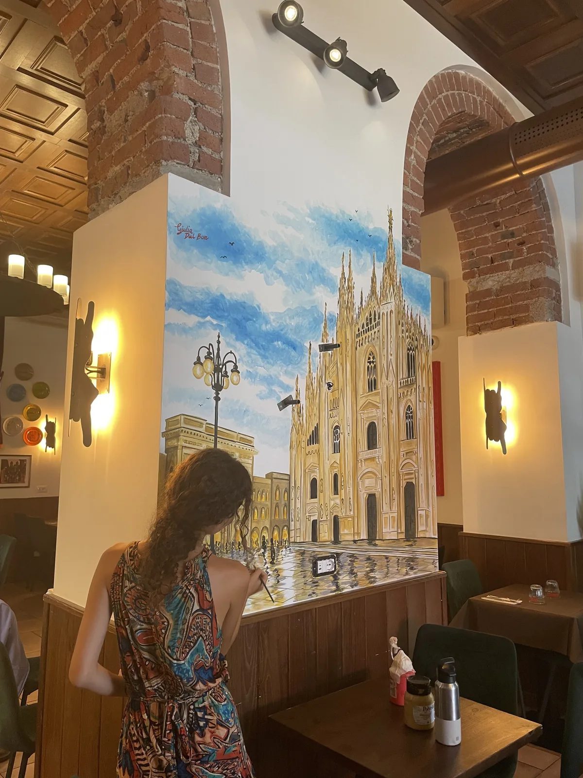 La parete è a colori, e Giulia Dal Bon dipinge i riflessi della piazza ai piedi del Duomo, sopra i tavoli del ristorante.