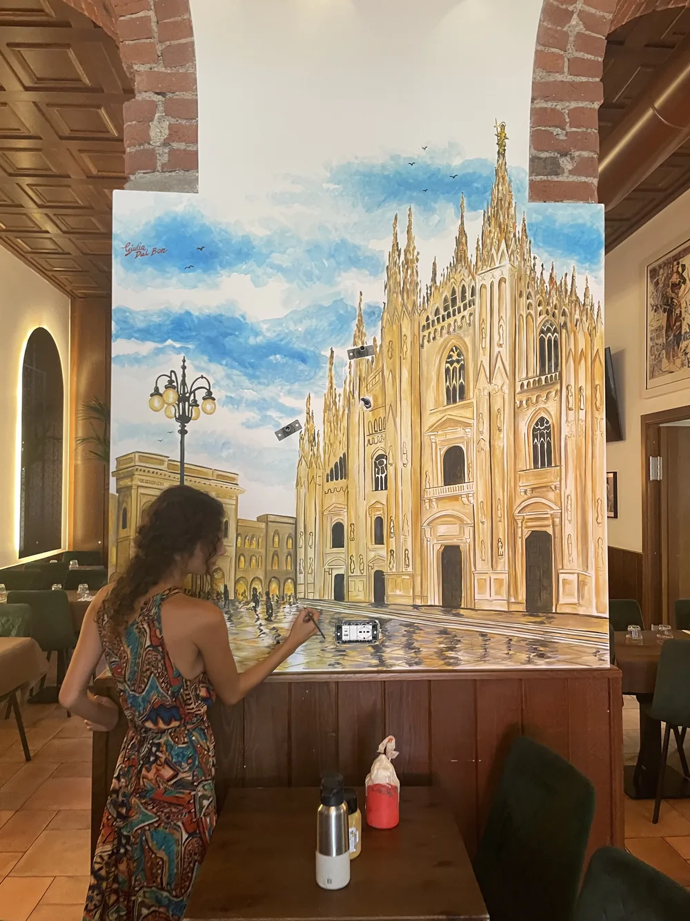 Giulia Dal Bon dipinge a pennello il murale del Duomo di Milano sotto un grande cielo azzurro, sulla parete di un ristorante.