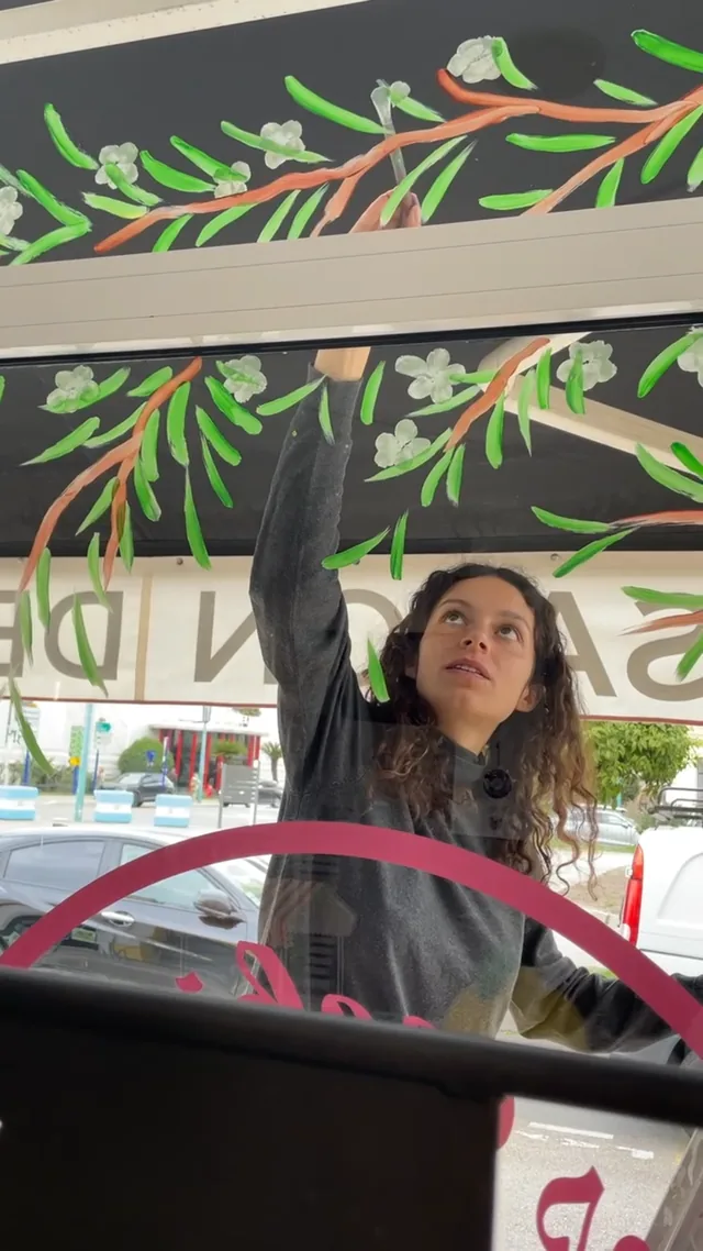 Derrière la vitre, Giulia Dal Bon peint des branches fleuries sur le haut d’une vitrine.